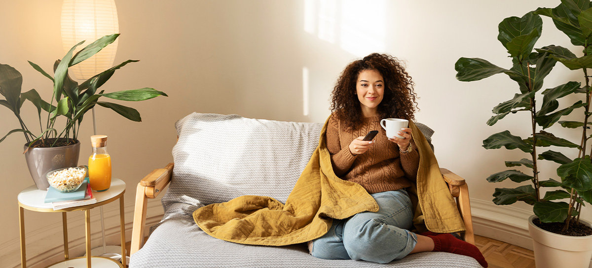 Woman sitting on couch in living room while holding remote, looking cozy.